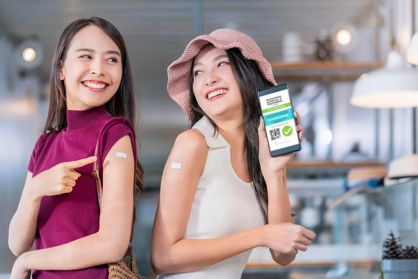 Весёлая улыбающаяся азиатская девушка-подросток жестом указывает на бренд вакцины от COVID‑19 и на экране смартфона показывает цифровой паспорт вакцинации после прививки; концепция профилактики здоровья.